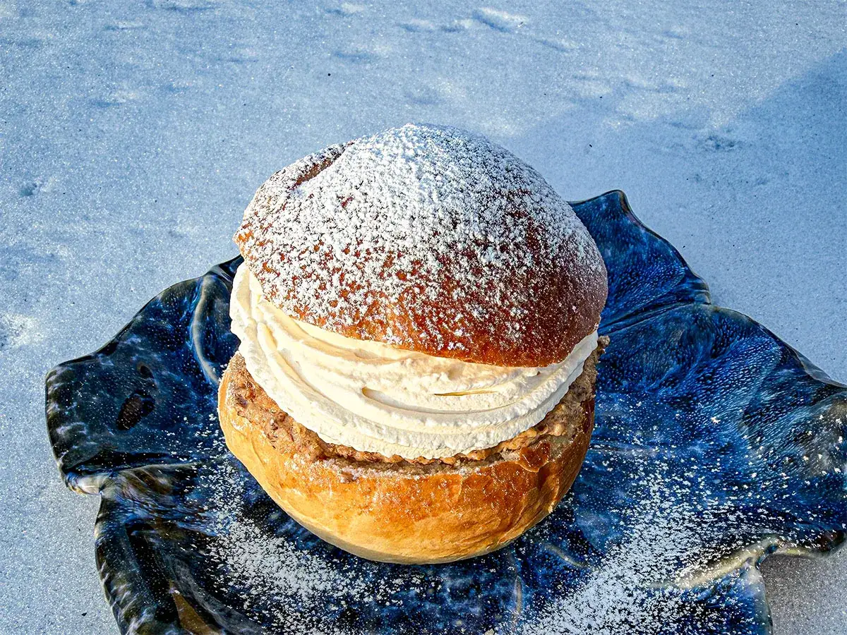 En semla med rikligt med gr&auml;dde och mandelmassa till semlor, pudrad med florsocker, ligger p&aring; en bl&aring;, v&aring;gig tallrik i sn&ouml;n.