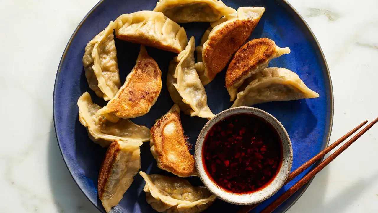 En bl&aring; tallrik med gyllene dumplings i buljong, serverade med en sk&aring;l med chilis&aring;s och &auml;tpinnar.