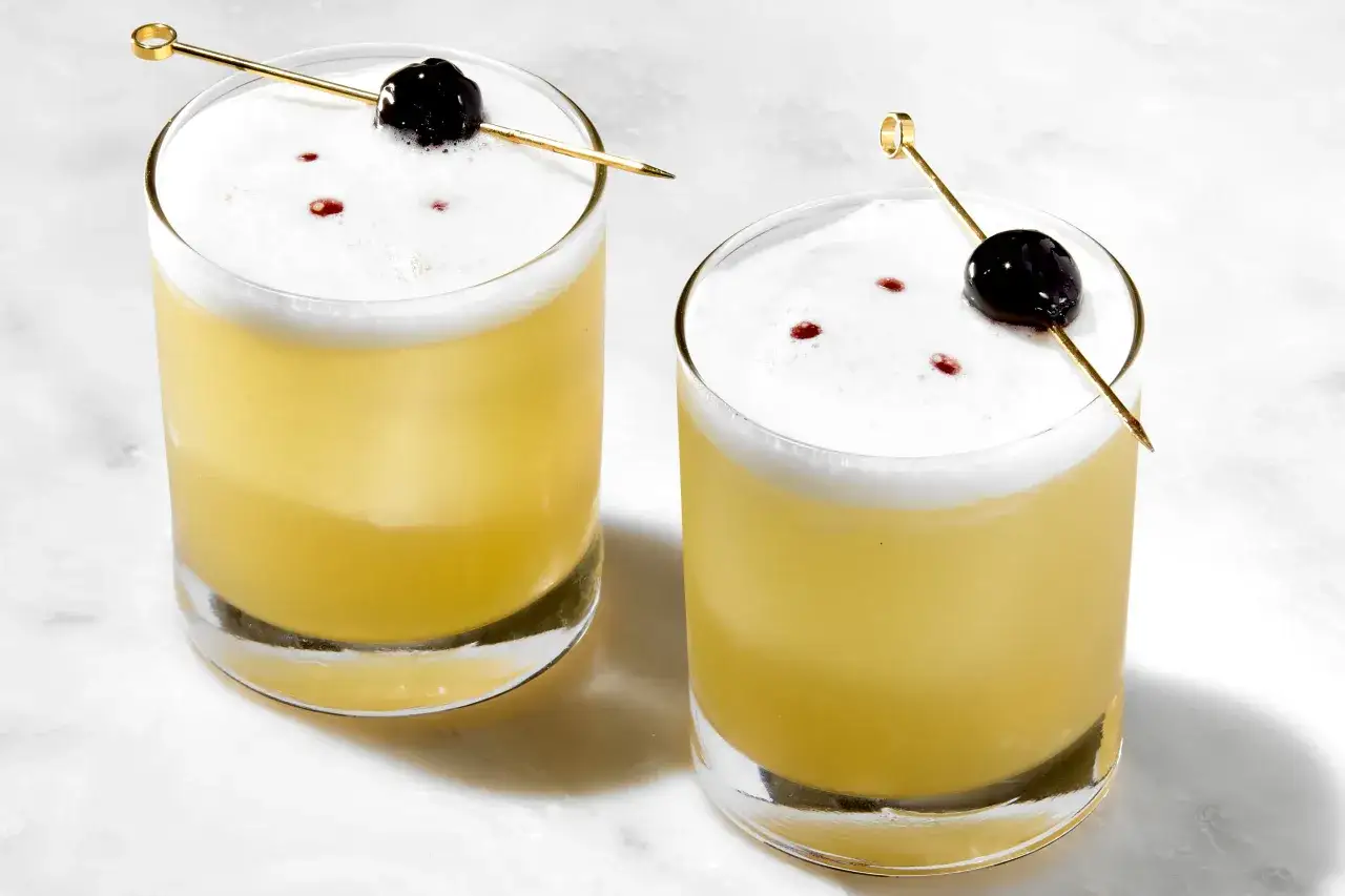 Tv&aring; glas med gyllene whiskey sour, toppade med vitt skum, en k&ouml;rsb&auml;rsgarnityr och en guldig cocktailpinne.