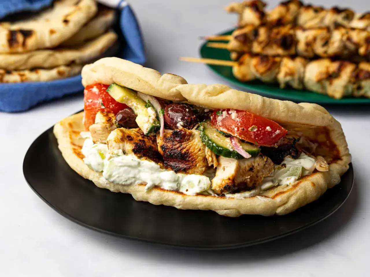 En saftig souvlaki kyckling-gyros med tzatziki, tomat, gurka och oliver i ett mjukt pitabr&ouml;d. I bakgrunden syns fler grillspett.