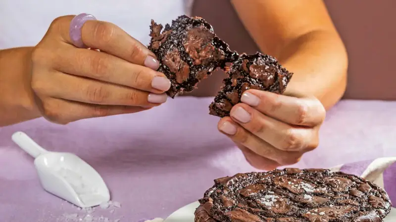 Händerna bryter isär en saftig chokladkaka, strödd med flingsalt. En hel kaka ligger på en tallrik.